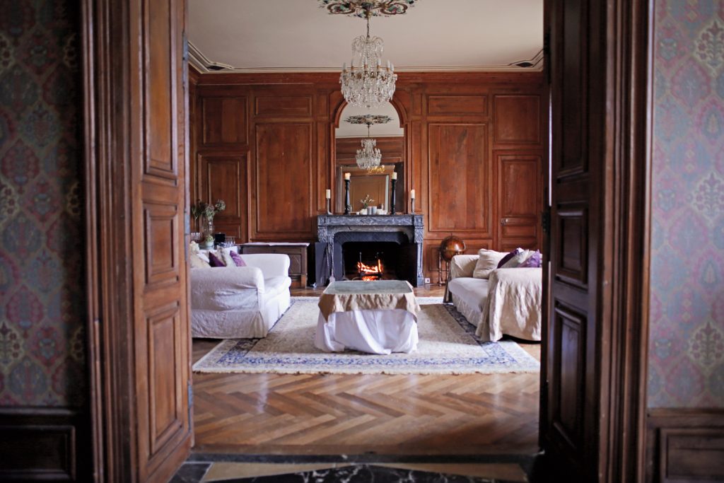 Elegant living room with a wooden interior, featuring a vintage chandelier and ornate ceiling. A fireplace with lit fire is centered on a dark marble wall. White sofas and chairs surround a coffee table on a patterned rug, viewed through open doors.