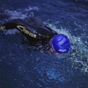 Person swimming in a pool, wearing a blue swimming cap and a black wetsuit with yellow lettering. They are doing a front crawl stroke, causing splashes in the dark water around them.