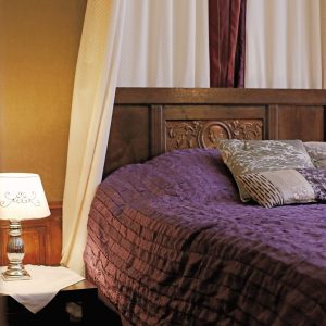 A cozy bedroom features a dark wooden bed with a purple quilted bedspread and patterned pillows. A nightstand beside the bed holds an elegant lamp with a decorative shade, casting a warm glow in the room.