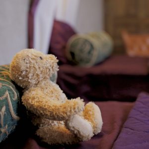A plush teddy bear sits leaning against an ornate green and gold pillow on a bed with purple bedding. In the background, there are more similar pillows and a wooden door, creating a cozy atmosphere.