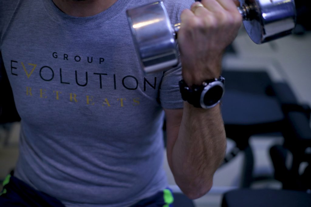 A person in a gray shirt with Group Evolution Retreats lifts a dumbbell with their right hand. They are wearing a watch on their wrist. The background shows gym equipment.