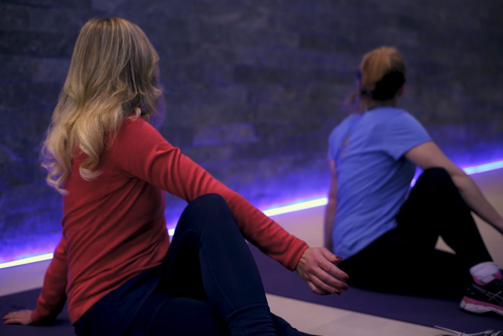 Two women sitting on yoga mats performing a seated twist pose. The woman in the foreground has long blonde hair and wears a red top, while the woman in the background has a ponytail and wears a blue shirt. A purple light illuminates the wall behind them.