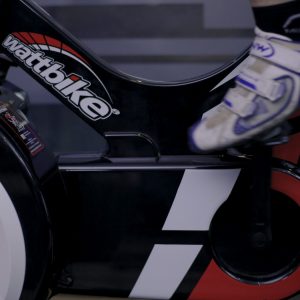 Close-up of a person using a Wattbike exercise bike. The focus is on the pedal area, with the persons foot in a cycling shoe secured in the pedal. The black bike features red and white accents.