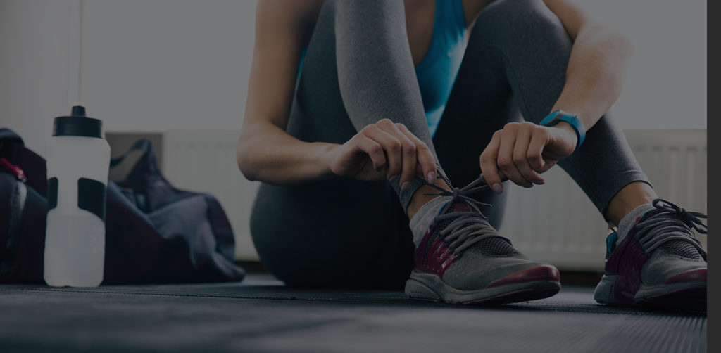 A person in athletic clothing sits on a gym floor, tying the laces of their sneakers. A water bottle and gym bag are nearby. The person is wearing a blue fitness tracker and gray leggings.