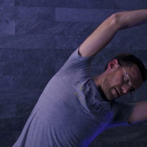 A man wearing glasses and a gray T-shirt with text is stretching with one arm over his head against a dark, textured wall.