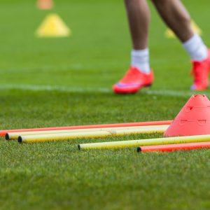 A person wearing bright pink athletic shoes runs across a grassy field, surrounded by several yellow training poles and an orange Sports movement screen cone scattered among the grass, while blurred Sports movement screen cones appear in the background.