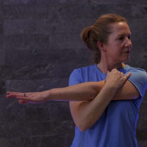 A woman with light brown hair in a ponytail stretches her right arm across her chest, supported by her left hand, while wearing a blue t-shirt against a dark stone wall backdrop. This image showcases the Swimming Technique Assessment product.