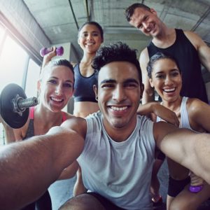 A group of five people in workout attire pose happily for a selfie in a gym. They are holding dumbbells, and natural light streams in from large windows behind them, highlighting their energetic and cheerful expressions.