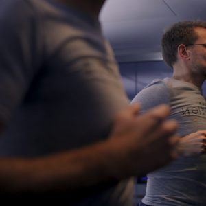 A man wearing glasses and a gray T-shirt is jogging on a treadmill, reflected in a mirror. The background is blurred, focusing on his concentrated expression and exercise posture.