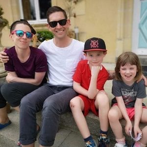 A family of four people sitting on outdoor steps. Two adults, both wearing sunglasses, are sitting on either side of two children. One child wears a black cap, red shirt, and red shorts. The other child wears a grey shirt and shorts. They all smile at the camera.