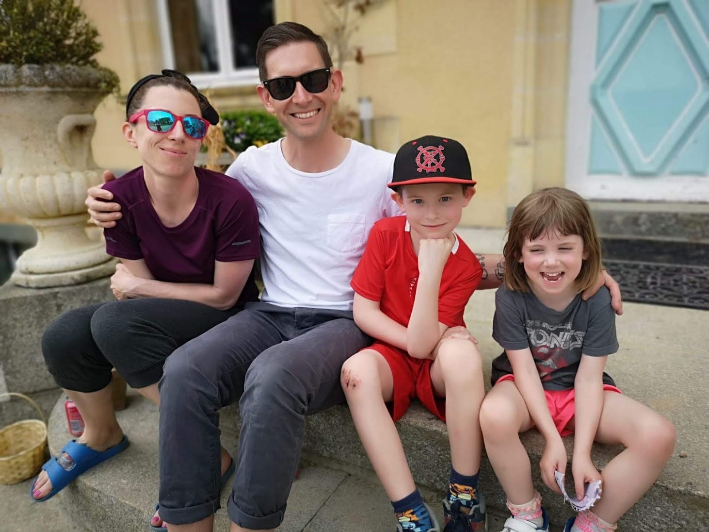 A family of four people sitting on outdoor steps. Two adults, both wearing sunglasses, are sitting on either side of two children. One child wears a black cap, red shirt, and red shorts. The other child wears a grey shirt and shorts. They all smile at the camera.