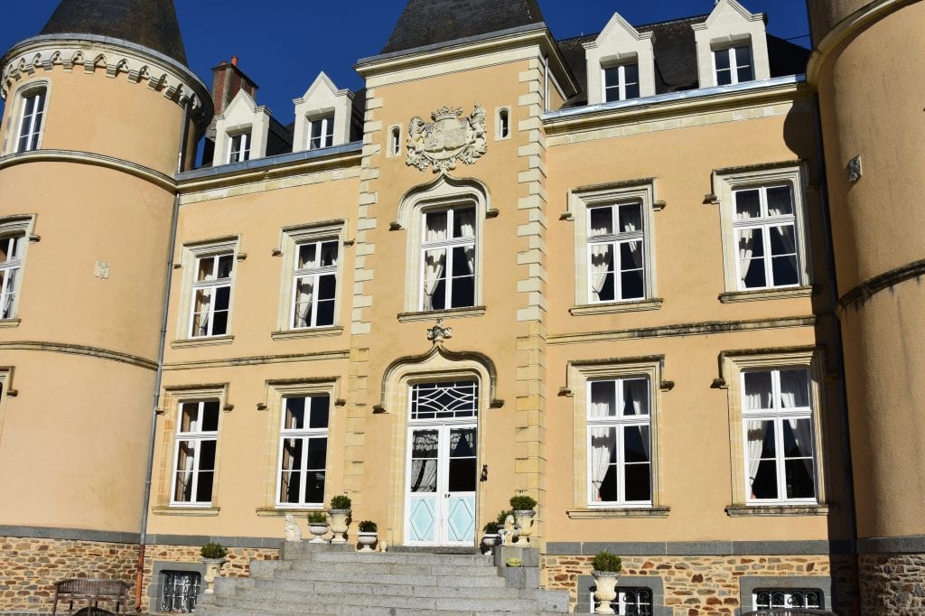 A grand, beige chateau with an ornate facade, featuring tall windows, a decorative crest above the entrance, and two round towers on either side. The building is set against a clear blue sky. Grey steps lead up to the door.