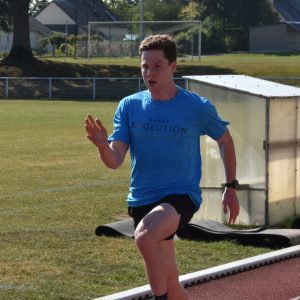 A person is running on an outdoor track wearing a blue Group Evolution shirt and black shorts. They are mid-stride, with a focused expression, and there are trees and buildings in the background.