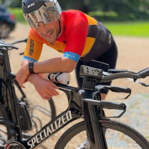 A cyclist in a red and orange jersey on the Going Long camp, wearing a reflective helmet and visor. Another bike is partially visible against the backdrop of a gravel path and blurred greenery.