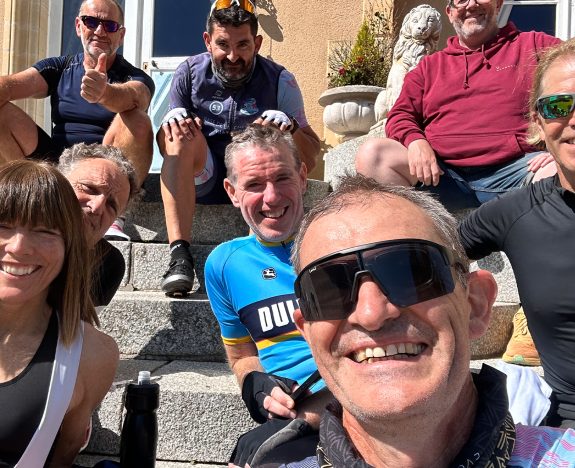 Cyclists enjoying a sunny day at the Torrelominos GB Age Group Camp, sitting on stone steps, wearing cycling gear and sunglasses. One in a red hoodie gives a thumbs-up while another captures the moment with a group selfie.