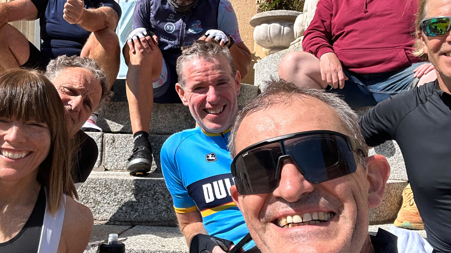 Cyclists enjoying a sunny day at the Torrelominos GB Age Group Camp, sitting on stone steps, wearing cycling gear and sunglasses. One in a red hoodie gives a thumbs-up while another captures the moment with a group selfie.