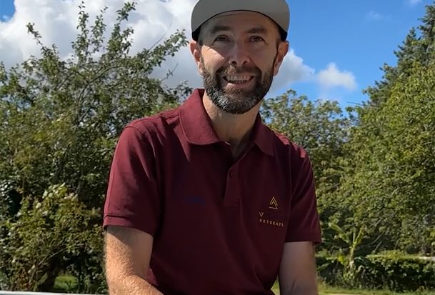 Joel Filliol, wearing a maroon polo shirt and a black cap, sits on a low wall by the pool at the Joel Filliol Retreat, smiling at the camera against a backdrop of trees and a blue sky with clouds.