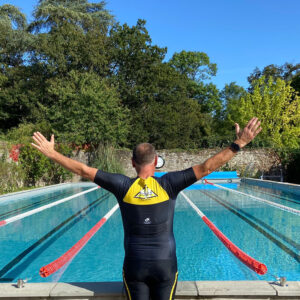 At Craig's Memorial Ride, a man in a wetsuit stands poolside with arms raised, preparing to dive in against a backdrop of greenery and blue sky during the endurance sports festival.