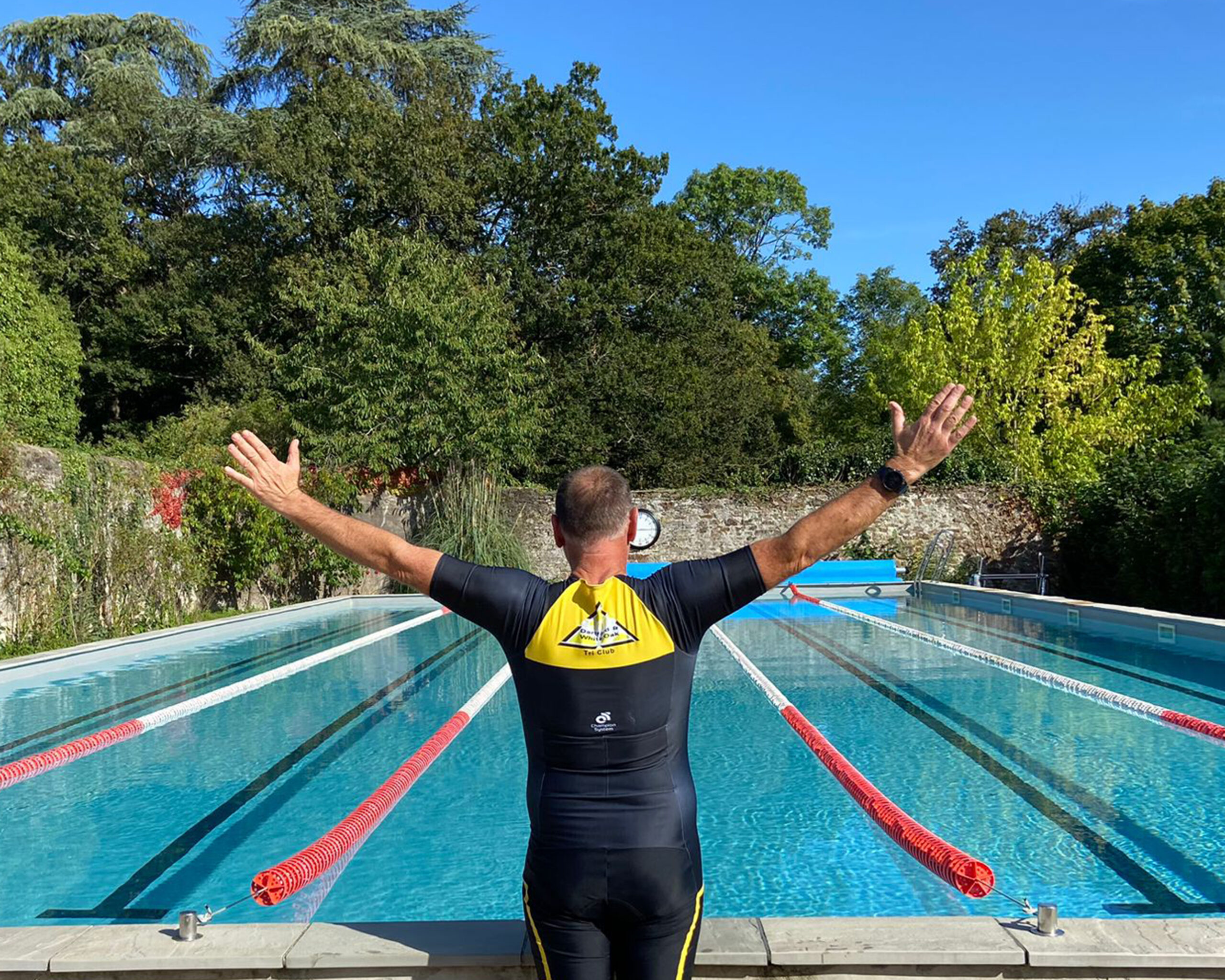 At Craig's Memorial Ride, a man in a wetsuit stands poolside with arms raised, preparing to dive in against a backdrop of greenery and blue sky during the endurance sports festival.