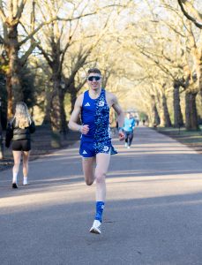 A man in blue athletic gear and sunglasses runs on a sunny, tree-lined park path, capturing the energetic vibe of Run Well with Nick Bester, as others walk and jog in the background.