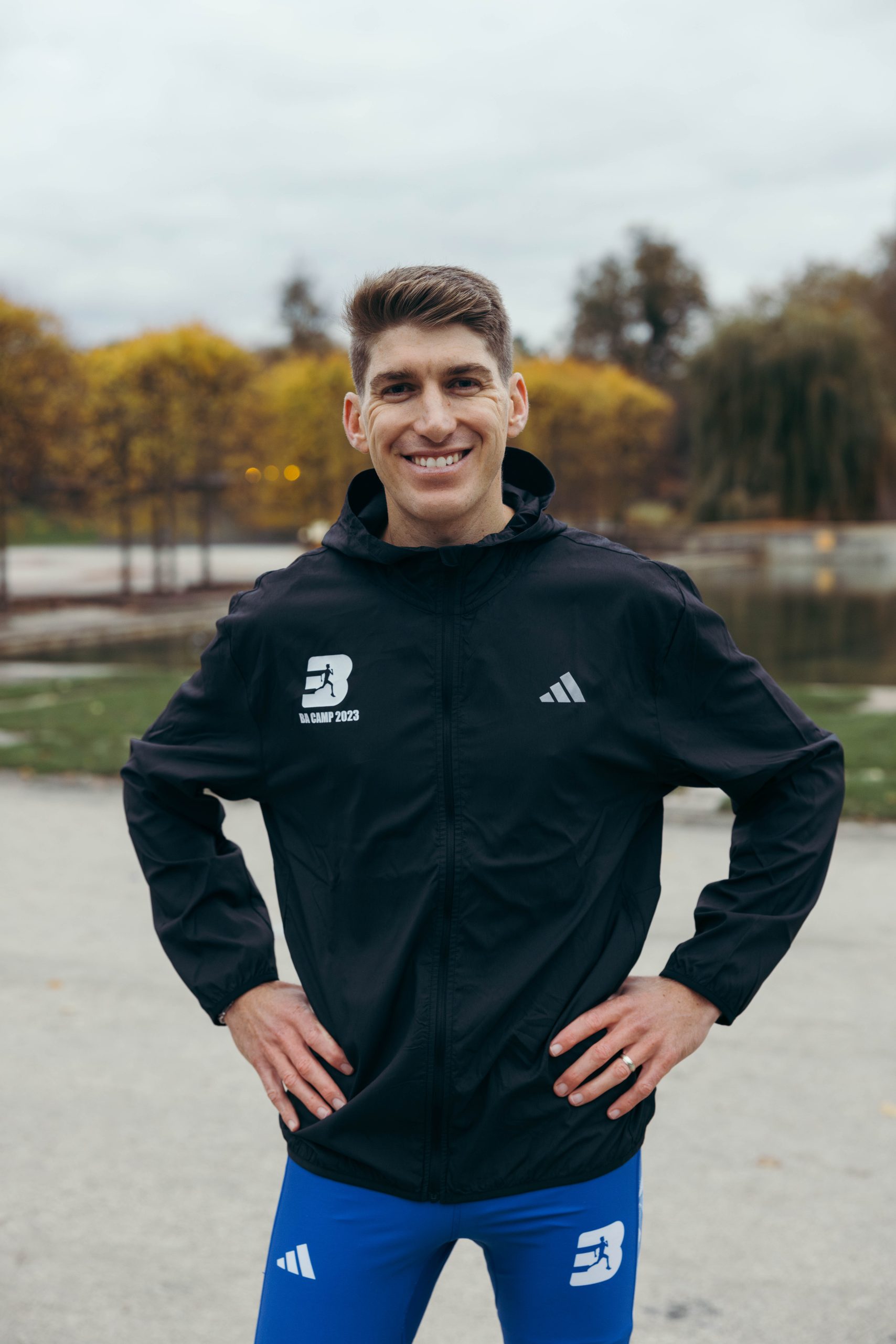 231110 - Best Athletics-44 A smiling man stands outdoors with his hands on his hips, wearing a black sports jacket and blue athletic pants—ready for a run training retreat with Nick Bester. Autumn trees and a pond provide a scenic backdrop.