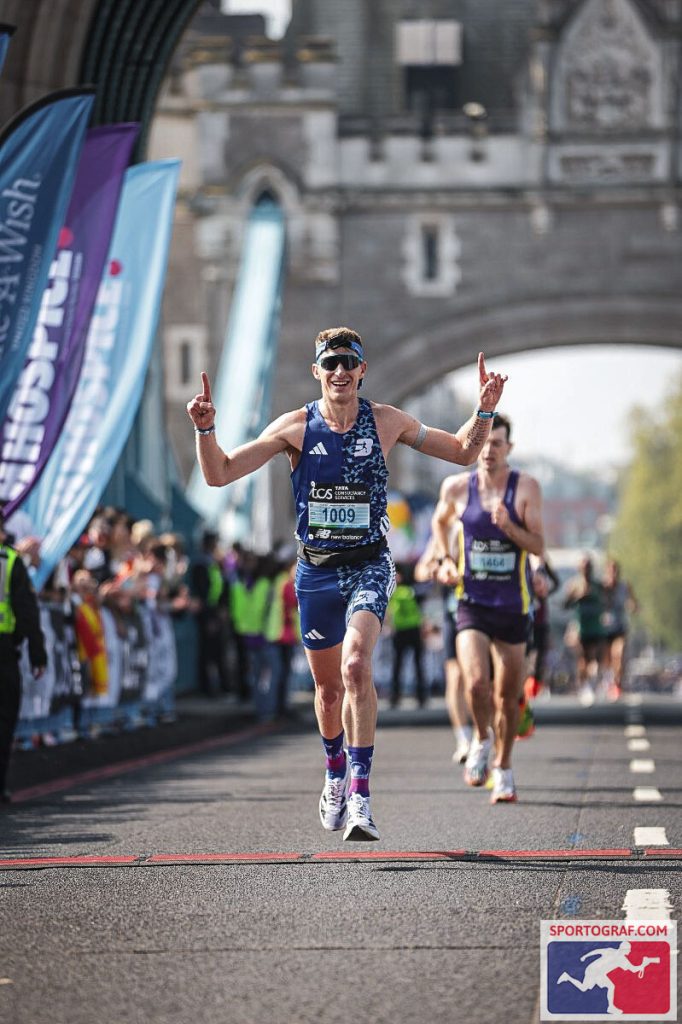 A marathon runner in sunglasses and a blue patterned outfit smiles and raises both hands in celebration on a city street, possibly inspired by Run Well with Nick Bester, with spectators and a historic stone bridge in the background.