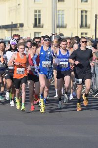 A group of runners in athletic gear, led by Best Athletics head coach Nick Bester, participate in a road race on a sunny day in an urban area, with race numbers pinned to their shirts and spectators cheering from the sidewalks.