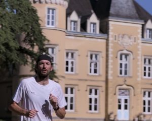 A man in a white shirt and black cap is running outdoors in front of a large, ornate yellow building—perfect inspiration for a run training retreat with Best Athletics head coach Nick Bester.