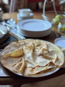 A plate of thin, golden crepes sits on a wooden table set for a meal at a glorious chateau in the Loire, with stacked white plates, cups, cutlery, and a fruit basket visible in the background.