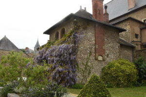 A stone building with red brick chimneys is covered in purple wisteria flowers, surrounded by green bushes and a garden. The sky is overcast and other old buildings are visible in the background.