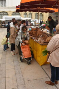 People shop at an outdoor market stall with a yellow table, selling brown packaged goods. A woman with a shopping trolley stands at the counter, while others wait in line. Red umbrellas provide shade.