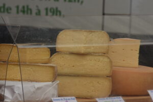 Several large wheels and blocks of yellowish cheese are stacked behind a glass display at a market, with some cheese labels visible and green text blurred in the background.