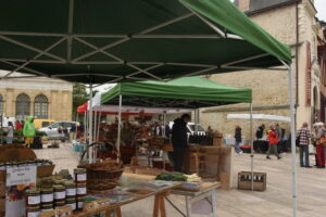 Outdoor market scene with green canopy tents, tables displaying jars and baskets, and people browsing and walking among various stalls in a town square with old stone buildings in the background.