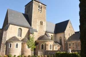 A historic stone church with a tall clock tower, arched windows, and a dark slate roof, surrounded by greenery and trees under a clear blue sky.