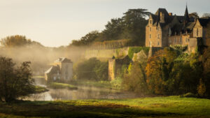 A historic stone castle sits atop a green hill, surrounded by autumn trees and morning mist, overlooking a calm river and smaller stone buildings below. Sunlight highlights the scene, creating a peaceful, dreamy atmosphere.