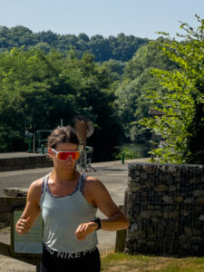 A woman wearing sunglasses and a tank top jogs outdoors on a sunny day, surrounded by greenery and trees, with a river, hills, and the silhouette of a glorious chateau in the Loire visible in the background.