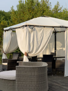 A wicker patio set with chairs and a round ottoman sits under a white canopy tent with draped curtains, surrounded by greenery on a wooden deck—evoking the charm of a glorious chateau in the Loire.