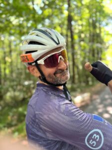 A man wearing a cycling helmet, sunglasses, and a purple cycling jersey smiles and gives a thumbs-up on a sunlit forested path near the glorious chateaux in the Loire.