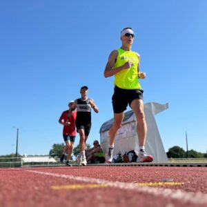 Four runners compete on a red track under a clear blue sky, with a Glorious Chateau in the Loire visible near the white structure. The front runner wears a neon yellow shirt and sunglasses. The photo is taken from a low angle.