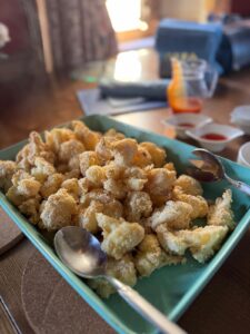 A rectangular teal dish filled with golden-brown, crispy breaded cauliflower florets, with two metal serving spoons—perfect for savoring at a Glorious Chateau in the Loire. In the background are small bowls of dipping sauces and a jug with red liquid.