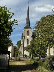 A stone church with a tall, pointed steeple and clock on its tower stands among trees, with sunlight casting shadows on the ground. A glass bus shelter and parked cars are visible nearby.