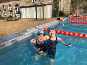 Two smiling girls in swimsuits and swim caps stand together in an outdoor swimming pool, holding goggles. The pool has lane dividers, and a stone building with arched windows is visible in the background.