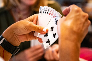 A person holds a fan of playing cards, mostly clubs and one heart, while another person sits across the table with blurred hands and face in the background. The focus is on the cards and hands.