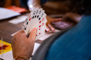 A person holds a fan of playing cards, including the 2, 5, 6, 9, and 10 of clubs, and the 10 of hearts, with another person and cards visible in the blurred background.