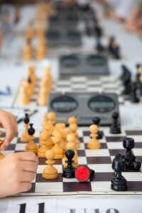 Close-up of a chess game in progress, with a childs hand moving a piece. Multiple chessboards and digital timers are visible in the background, and a black chess piece with a red sticker is tipped over on the board.