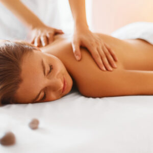 A woman lies face down on a massage table, smiling with her eyes closed, while receiving a back massage from a therapist. White towels and massage stones are visible in the soft, relaxing setting.