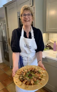 A smiling woman wearing glasses, a white apron, and a dark shirt holds a large platter of skewered food garnished with herbs in a kitchen setting.