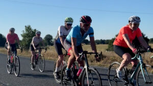 A group of cyclists ride on a rural road in sunny weather, surrounded by fields and trees. The lead cyclist wears a blue jersey, while others wear various colors including orange, white, and pink.