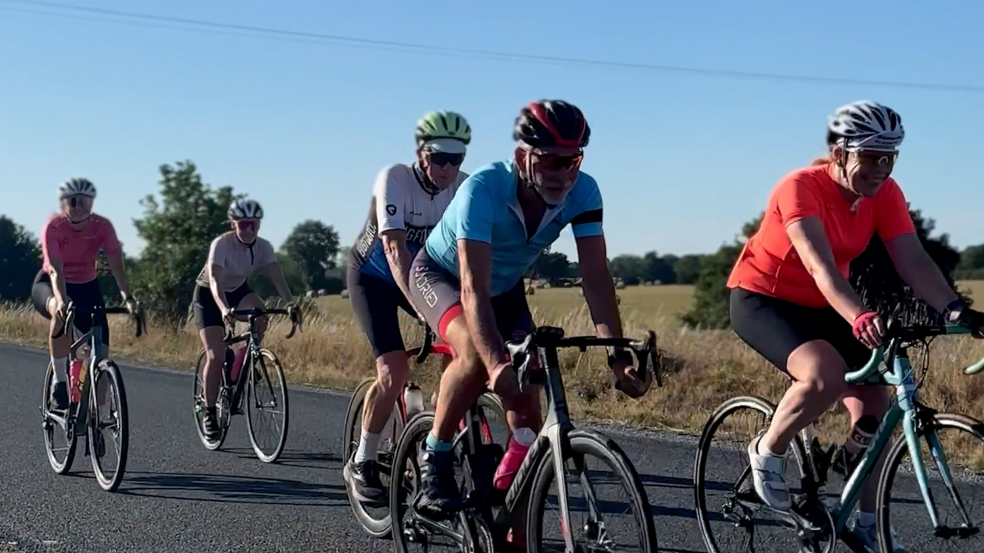 A group of cyclists ride on a rural road in sunny weather, surrounded by fields and trees. The lead cyclist wears a blue jersey, while others wear various colors including orange, white, and pink.