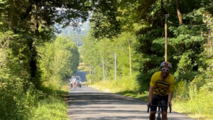 A cyclist in a yellow jersey rides uphill on a tree-lined road in sunlight, with several more cyclists visible in the distance behind him.